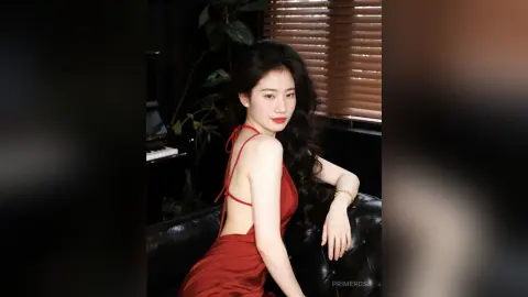 Elegant woman in a red satin slip dress sitting on a black leather sofa in a moody, dimly lit room.
