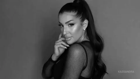 Glamorous woman in a black mesh top posing with a sleek high ponytail in a moody black and white studio portrait.