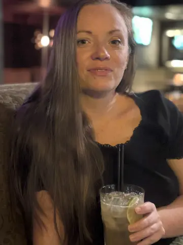 Smiling woman in a black top holding a refreshing iced cocktail in a dimly lit restaurant setting.