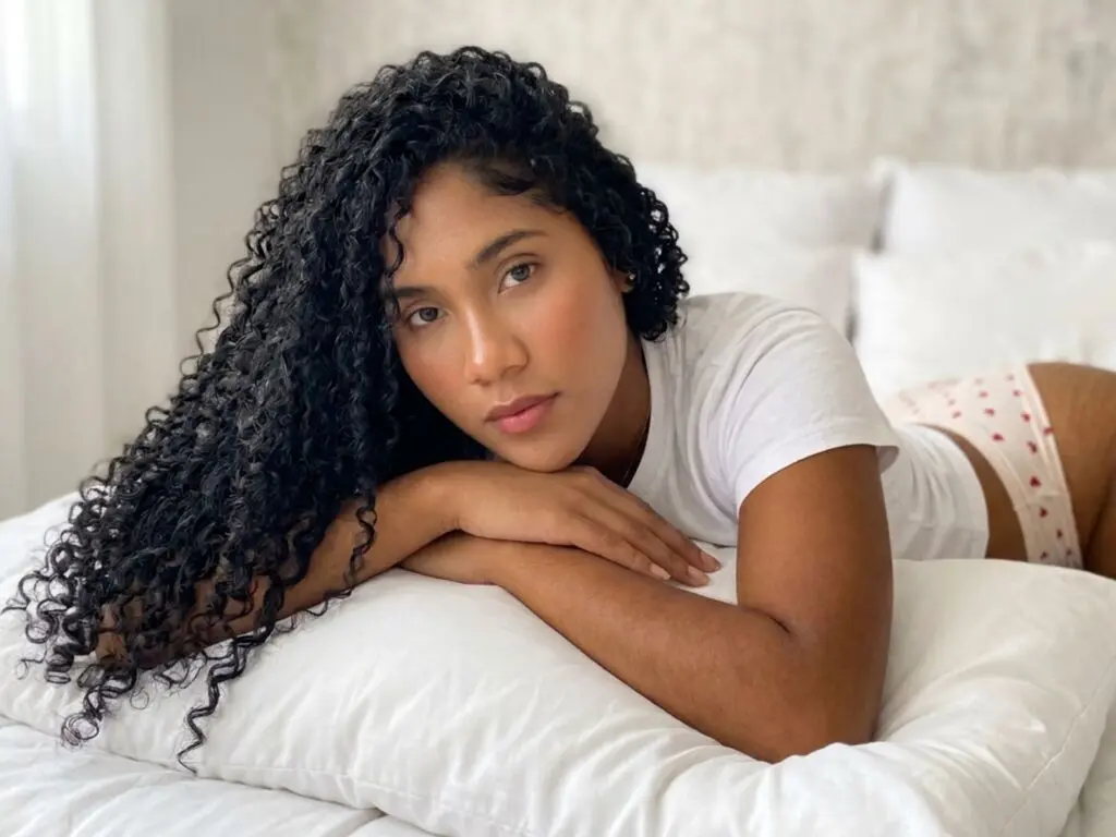 Young woman with long curly hair wearing a white t-shirt relaxing on a white bed in a bright, peaceful mood.