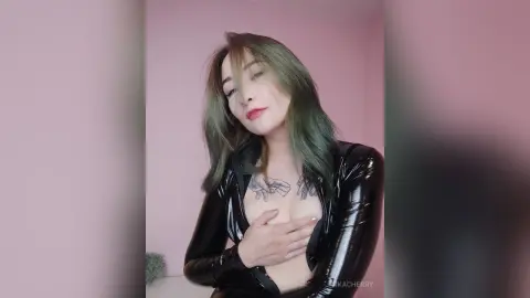 Young woman with long green hair wearing a black shiny latex bodysuit in a soft pink room.
