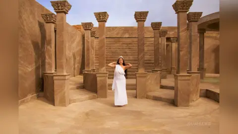 JuliahLewis stands in a white dress against a backdrop of tall stone columns and sand-colored walls.