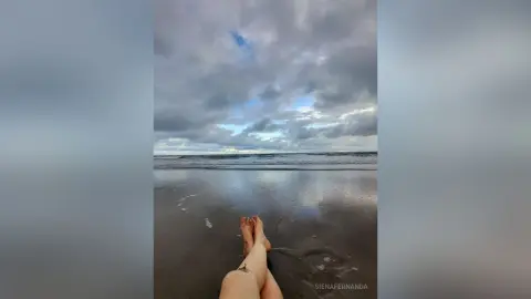Close-up of bare feet on a wet sandy beach under a moody, overcast cloudy sky at twilight.