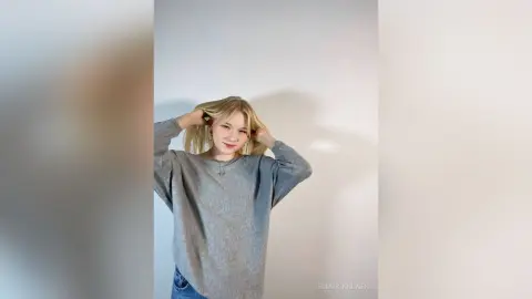 Smiling blonde woman in a cozy grey oversized sweater posing against a minimalist white background.