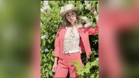 Young woman in a coral suit and white lace top posing amidst blooming white flowers in a sunny garden.