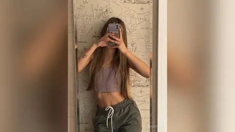 Young woman in a purple crop top and grey sweatpants taking a mirror selfie against a map wallpaper background.