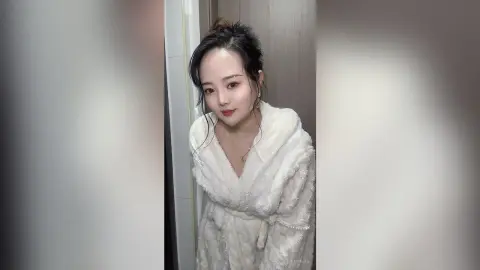 EnyaLin wears a white fluffy robe while looking at the camera with her dark hair tied up in a bun.
