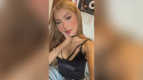 Young woman with winged eyeliner in a black tank top posing confidently in a cozy bedroom setting.