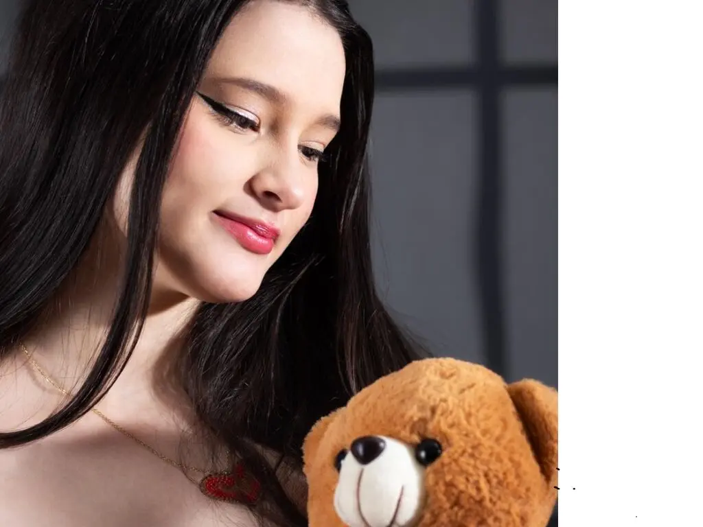 Smiling young woman with long dark hair holding a brown teddy bear in a soft, cozy indoor setting.