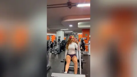 Blonde woman in white athletic top and purple shorts taking a selfie in a modern gym setting.