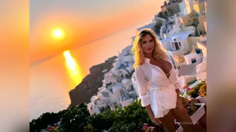 Blonde woman in a white breezy outfit enjoying a sunset view over Santorini coastal cliffs.