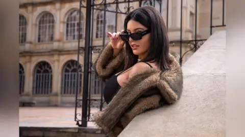 Stylish woman in a brown faux fur coat and sunglasses leaning against a stone wall in a classic courtyard.
