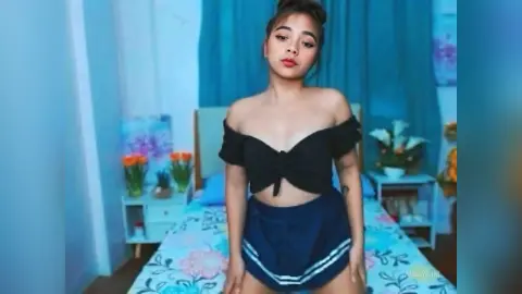 Young woman in a black off-the-shoulder crop top and navy shorts sitting on a bed in a bright blue room.