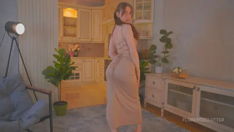 Young woman in a beige backless maxi dress posing in a cozy, warm-lit modern living room.