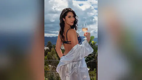 VeronicaValdez poses on a balcony wearing a blue bra and a white satin robe while holding a glass of champagne.