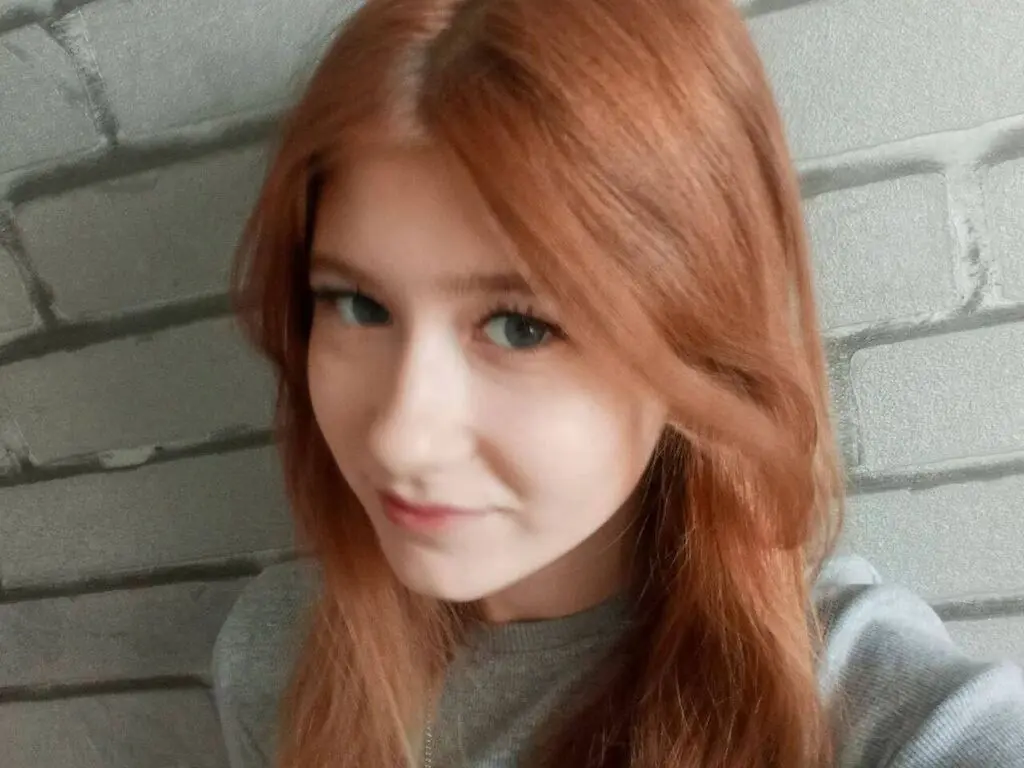 Smiling young woman with long red hair wearing a grey sweater against a grey brick wall background.