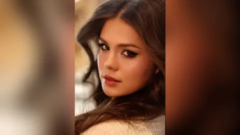DemetriceTaneja poses with a direct gaze, showing her long brown hair and heavy eye makeup against a soft, blurred background.