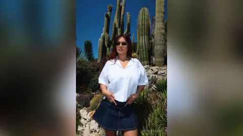 Stylish woman in a white button-down shirt and denim skirt posing in a sunny desert landscape with tall cacti.