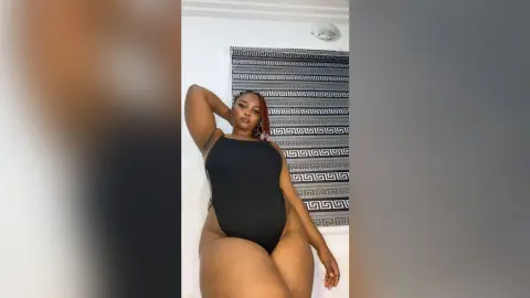 Confident woman in a black bodysuit posing against a patterned backdrop in a bright room.