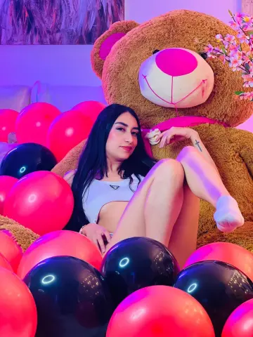 Young woman in a white crop top sitting by a giant teddy bear and red balloons in a dreamy pink setting.