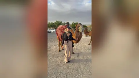 AlanaPearce stands outdoors in a black top and tan pants, posing next to a camel in a sandy desert setting.