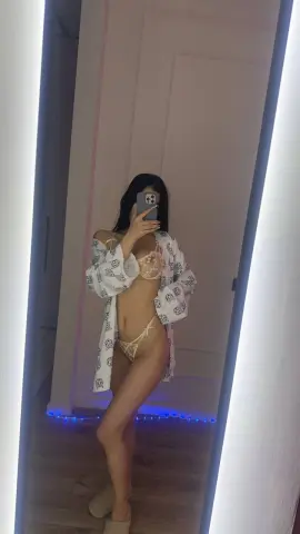 Young woman in beige lace lingerie and a white patterned robe posing for a mirror selfie in a modern room.