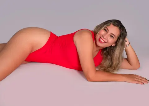 Smiling woman in a red bodysuit lying on a white studio background for a cheerful fashion portrait.