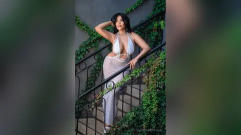 DayanaHille poses on a staircase wearing a white cut-out bodysuit with her hands behind her head against a grey wall and green ivy.