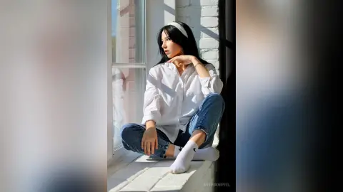 Melancholic young woman in white button-down shirt and jeans sitting by a sunny window.