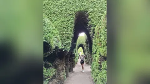 Woman in black outfit standing in a lush green hedge maze tunnel under bright daylight.