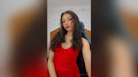 Young woman with curly hair wearing a red dress in a cozy bedroom setting.