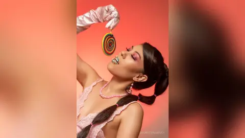 SamanthaHarvey holds a colorful swirl lollipop above her mouth while wearing white satin gloves and a pink lace top.