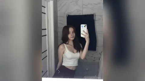 Young woman in a white ribbed tank top taking a mirror selfie in a modern marble bathroom.