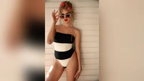 Stylish woman in a black and white striped swimsuit and sunglasses posing against a white paneled wall.