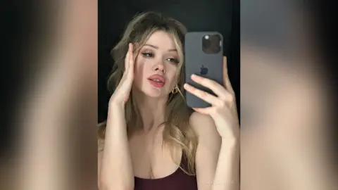 CassieArmas poses for a mirror selfie, touching her face and showing off her long blonde hair and cleavage in a dark tank top.