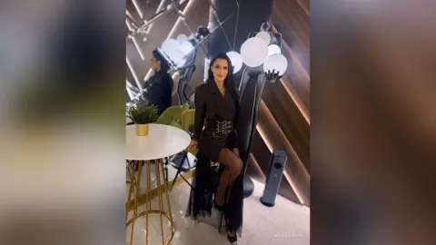 LeyaGrifin poses in a black outfit and sheer skirt while standing in a modern room with bright globe lights.