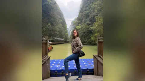 Smiling woman in a brown sweater and jeans standing on a wooden pier by a scenic river and lush mountains.