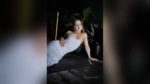AliceTucci poses on a dark floor wearing a white tube top, leaning back on one hand with her blonde hair down and gold jewelry.