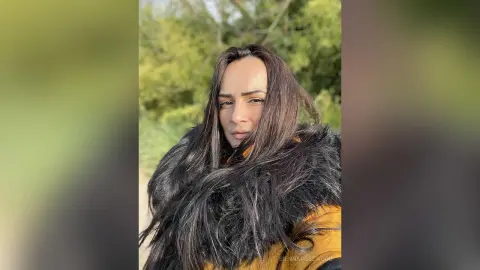 Woman with long dark hair wearing a cozy black faux fur coat in a sunny outdoor park setting.