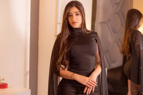 DulceMoretti poses in a black sleeveless turtleneck dress with a sheer cape, showing off her long brown hair and gold bracelet.