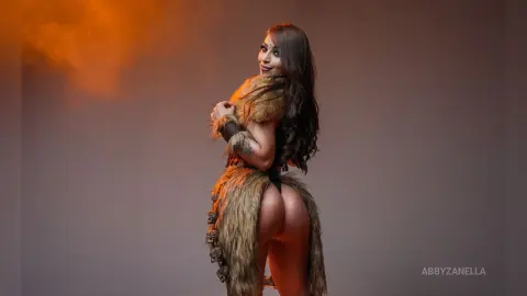 Smiling woman in a brown faux fur barbarian costume posing against a warm, moody studio background.