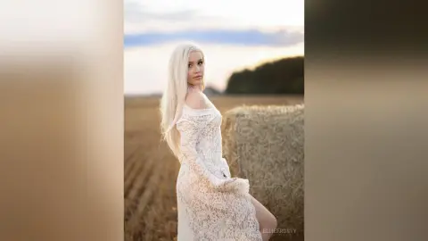Blonde woman in a white lace dress posing in a golden hay field during a dreamy sunset.