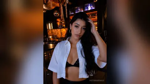 Young woman in a white button-down shirt and black bralette posing in a moody, dimly lit lounge setting.