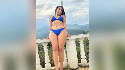 Smiling woman in a blue bikini posing on a white balcony overlooking a scenic mountain landscape.