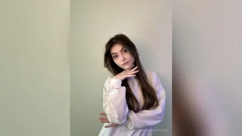 Young woman in a white long-sleeve sweatshirt posing thoughtfully against a neutral wall.