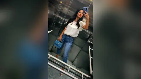 Stylish woman in a white ruffled blouse and blue jeans taking a mirror selfie in an elevator.