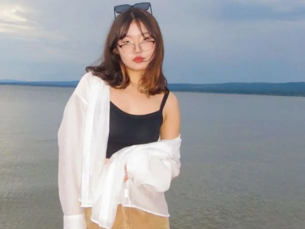 Young woman in a black crop top and white cardigan relaxing by the ocean at sunset.