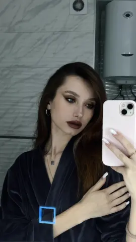 Young woman with long brown hair in a dark velvet robe taking a moody mirror selfie in a modern bathroom.