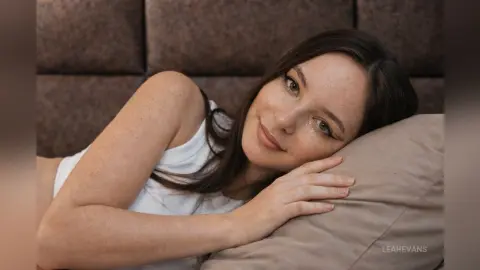 LeahEvans lies on a pillow in a white tank top, smiling at the camera with her long dark hair spread out.