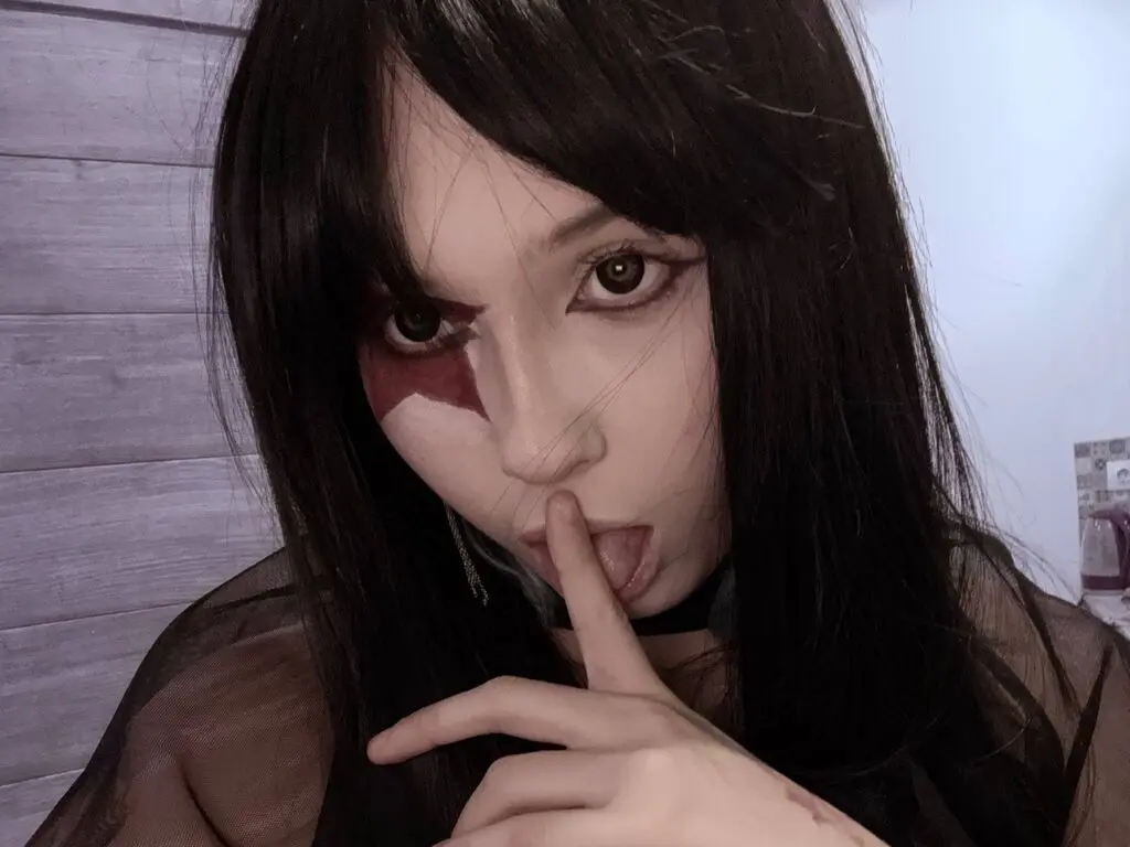 Woman with dark hair and dramatic eye makeup posing in a dark, moody setting.